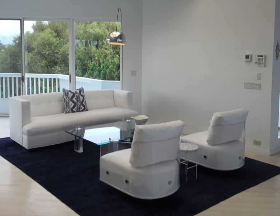 White sofa and two white chairs with acrylic arm-rests divided by clear acrylic coffee table sitting on blue area rug in all white walled room facing glass doors that open to white patio.